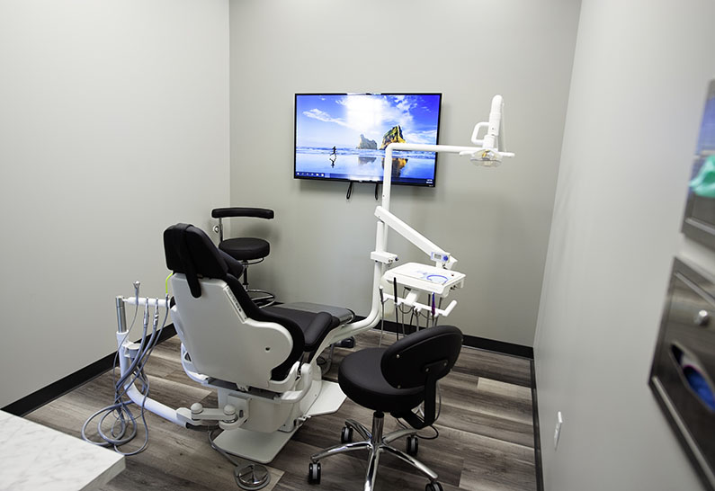 A dental exam room with a dental chair in the middle, an instrument tray to the right, and a tv on the wall