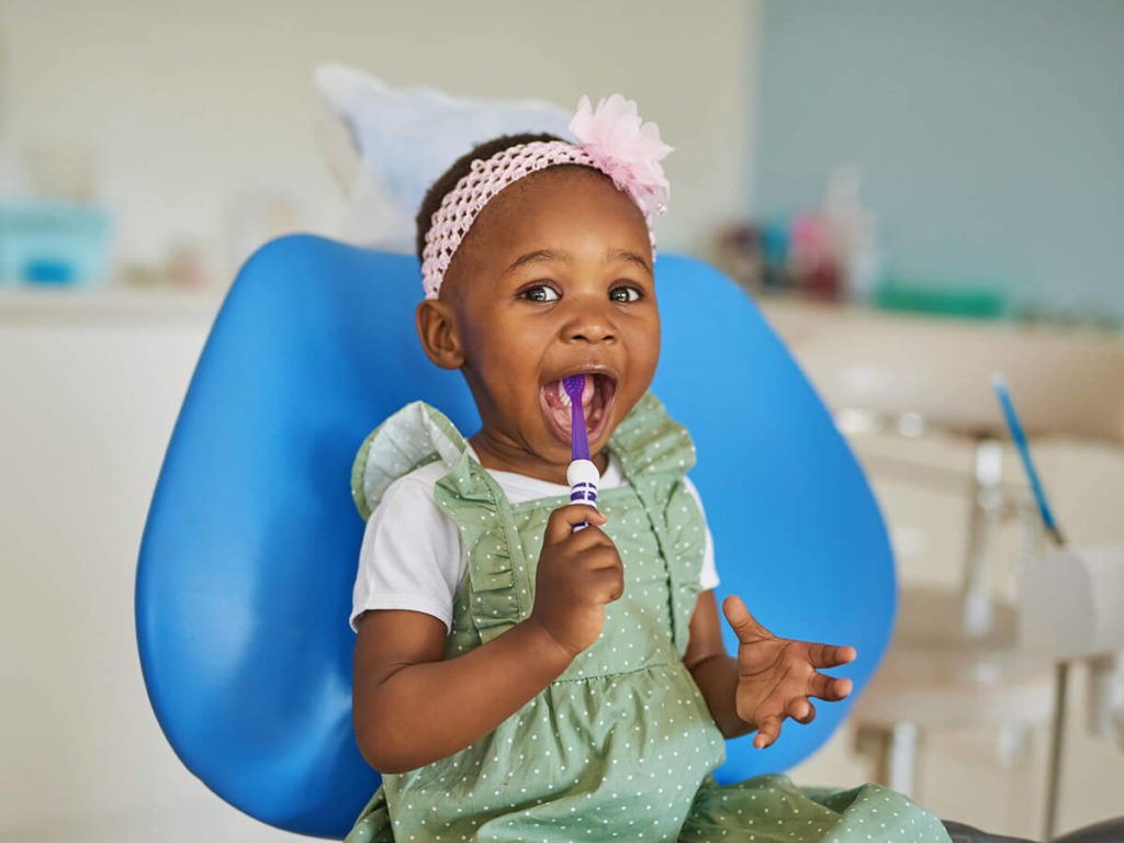 A young girl sits in a dental exam chair with a toothbrush in her mouth at a pediatric dentistry appointment