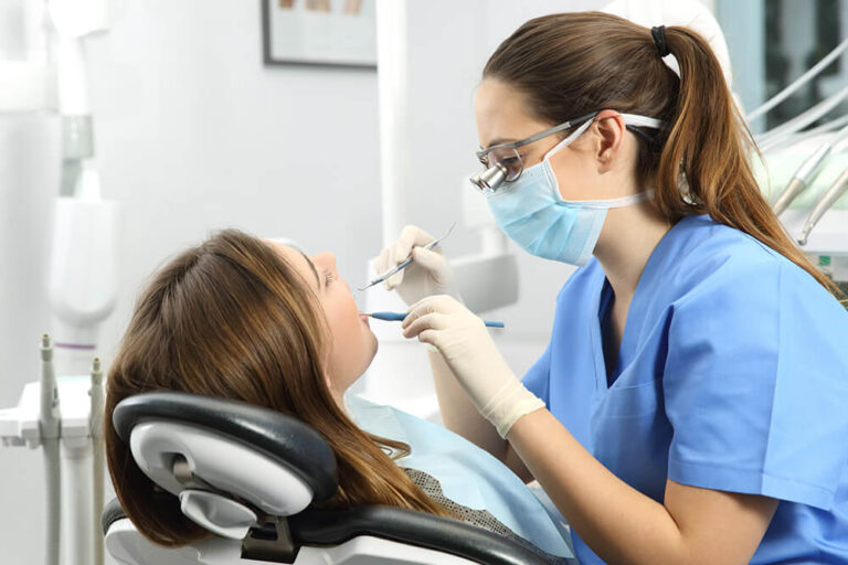 Dental hygienist in a mask and gloves performing a teeth cleaning on a patient in a dental chair.