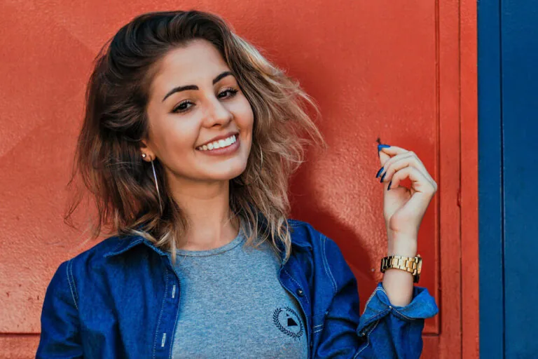 Young woman with wavy hair smiling confidently in front of a vibrant red background, wearing a denim jacket and a gold watch