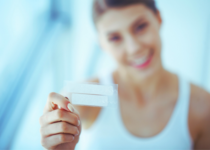 Smiling woman holding a teeth whitening strip in focus, with a blurred background.