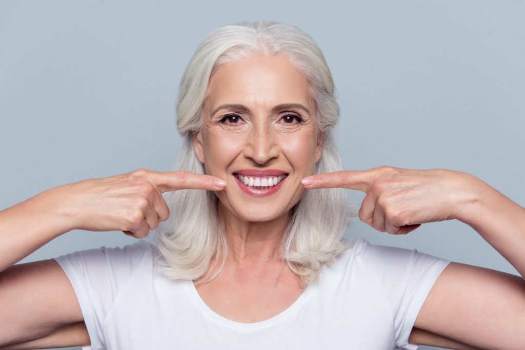 An older woman with grey hair pointing to her bright, white teeth