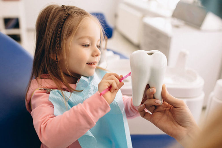 A young girl sits in a dental exam chair and brushes a model of a tooth that an out of view adult is holding