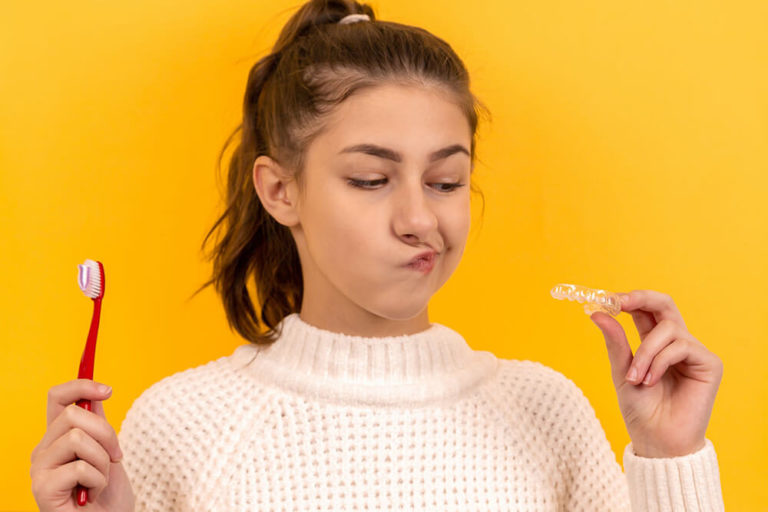 A young woman holds a toothbrush in one hand and a pair of Invisalign clear aligners in the other while making a skeptical face